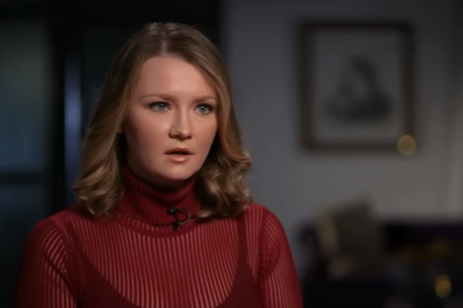 A woman with blonde curled hair wears a red sheer striped turtleneck sweater and looks ahead seriously during a seated interview. The background is softly blurred, showing a dim room with framed art and a couch, creating a focused and introspective atmosphere.