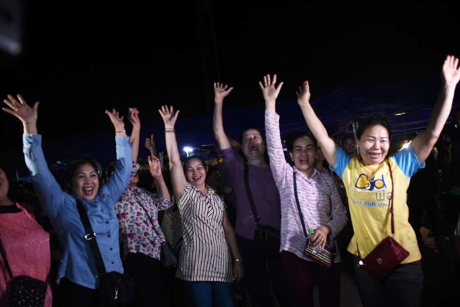 A group of women cheer joyfully with raised hands in a nighttime scene, celebrating the successful rescue of the trapped boys and coach from the Tham Luang cave. Their expressions reflect overwhelming relief and happiness, capturing the emotional climax of the rescue mission.