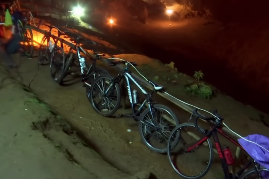 Several bicycles are parked in a row and secured to a rail outside a cave entrance at night, illuminated by artificial lighting. The scene suggests a pause or transition, possibly during a rescue or exploration effort, with the cave mouth visible in the dim background.