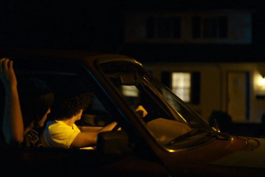 Two people sit in a parked car at night, with the driver looking toward a lit house across the street. The scene is dimly lit, creating a mysterious and contemplative atmosphere.