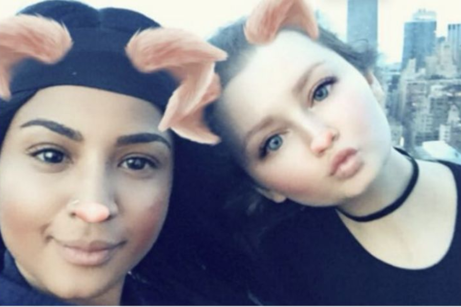 Two young women pose for a selfie using a playful animal face filter that adds pink ears and noses, giving them a cartoon-like appearance. The woman on the left wears a black headwrap and has a nose piercing, while the woman on the right puckers her lips and wears a black choker, with a city skyline visible in the background.
