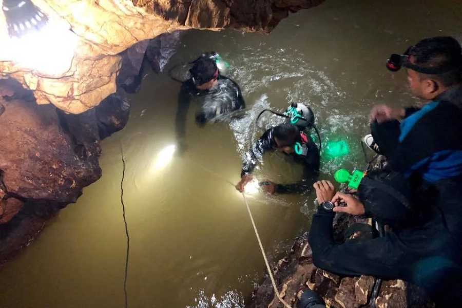 Divers in full scuba gear are seen entering a murky, narrow section of a cave, illuminated by headlamps and overhead lights. Two people observe and assist from the rocky ledge above, while one diver holds onto a guide rope as they begin their underwater navigation.
