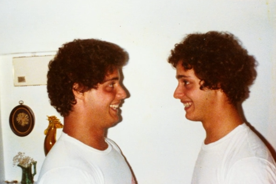 Two young men with matching curly hair and white shirts smile brightly at each other, standing face to face against a plain white wall in what appears to be a home setting.