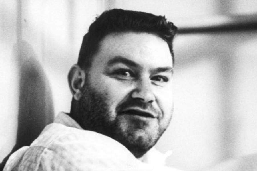 Black and white candid photo of a man with a trimmed beard and wavy hair, wearing a light quilted jacket. He is turning toward the camera with a slightly surprised or expressive look.
