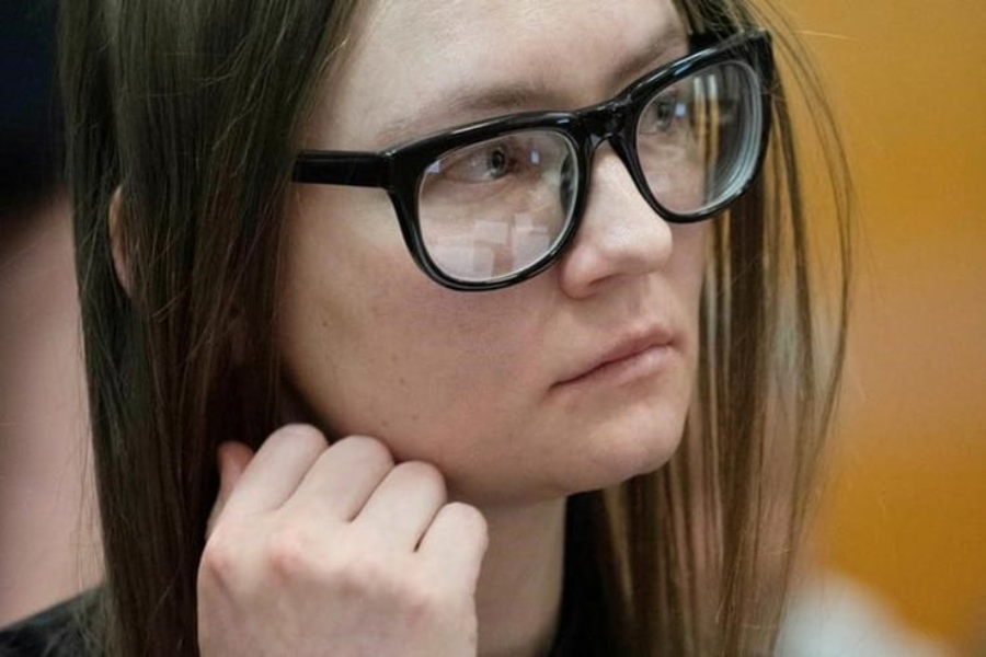 Close-up of a woman with straight brown hair and large black glasses, looking thoughtful and slightly tense as she rests her hand near her chin. The reflection of a courtroom is visible in her glasses, suggesting a legal setting.