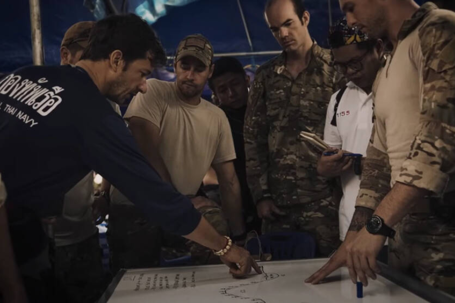 A group of rescue personnel, including military members in camouflage and a man in a Thai Navy shirt, gather around a whiteboard discussing a cave rescue plan. They appear focused and serious as one man points at a hand-drawn map on the board, outlining the operation strategy.