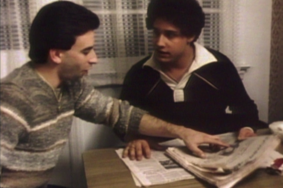 Two men sit at a kitchen table looking at a newspaper, with one pointing to an article and the other appearing engaged in the discussion. The scene feels candid and intimate, with warm lighting and casual 1980s-style clothing.