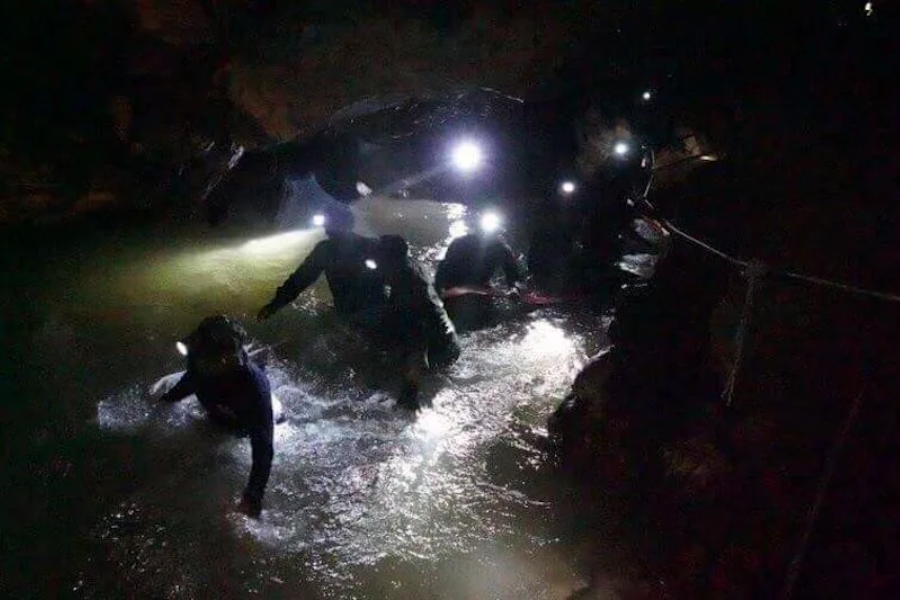 A rescue team wearing headlamps carefully wades through waist-deep water inside a dark, narrow cave. The beams of their lights reflect off the water’s surface, highlighting the treacherous, rocky environment as they follow a guide rope secured to the cave wall.