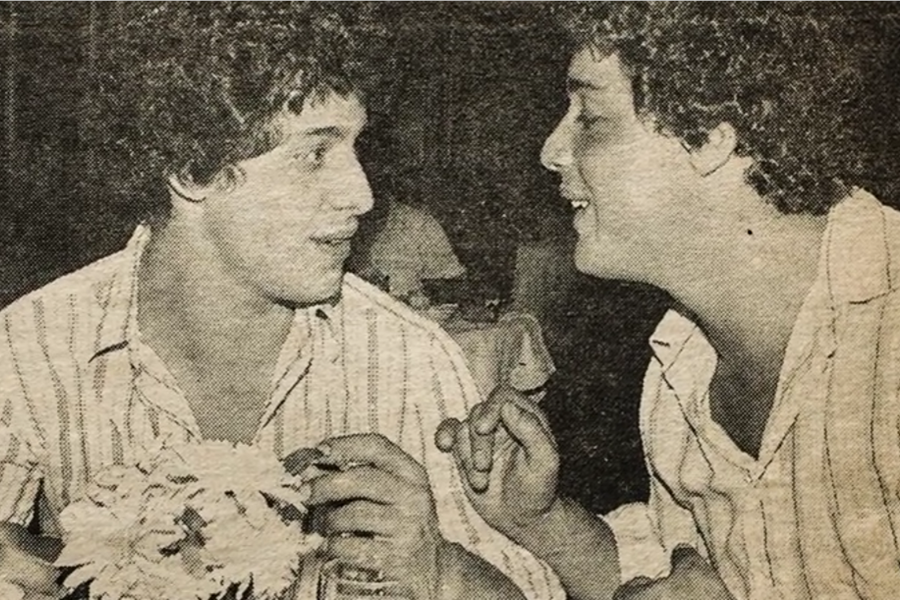 Vintage black-and-white photo of two young men wearing matching striped shirts, sitting close together in conversation. One holds a bouquet of flowers while the other gestures animatedly, both smiling and deeply engaged.