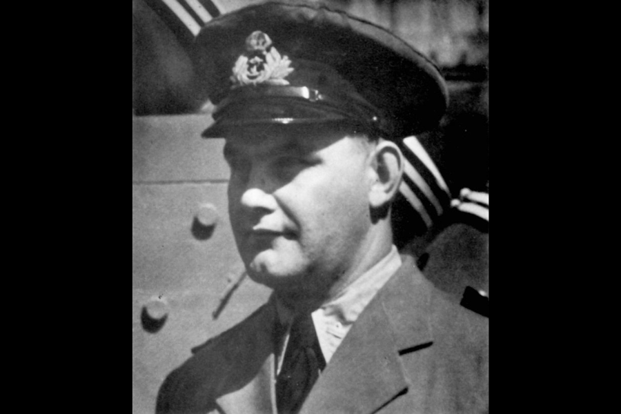 Black and white photo of a man in a military or naval officer’s uniform, including a peaked cap with an emblem. He is looking to the side, and the background includes a metal surface and another uniformed figure.