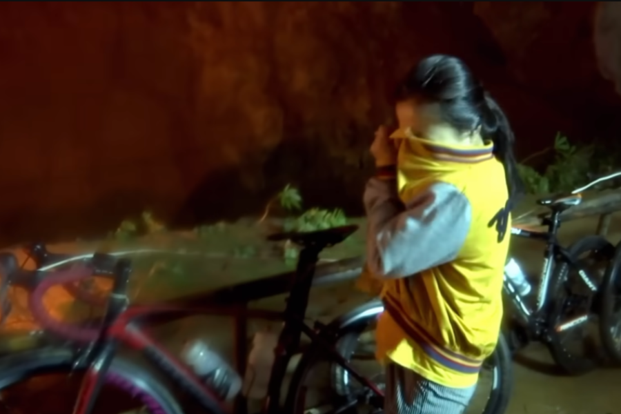 A young girl in a yellow and gray jacket stands beside a row of bicycles near a cave entrance at night, covering her face with her shirt in a gesture of worry or distress. The scene is dimly lit, with the cave’s earthy walls glowing under artificial lighting in the background.