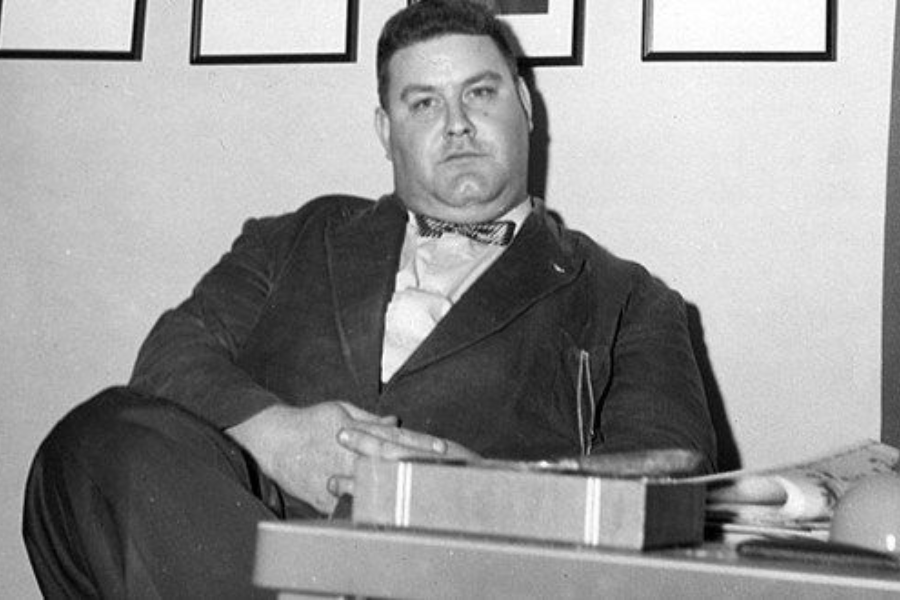 Black and white photo of a man sitting in an office-like setting, dressed in a suit with a bow tie. He has a serious expression, with his hands resting on his lap, and a desk with books and papers is visible in the foreground.