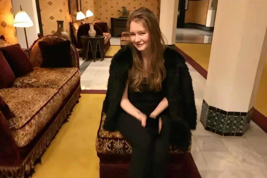 A woman sits on an ornate bench in a luxurious, warmly lit room with vintage-style velvet furniture and patterned decor. She wears a black outfit with a black fur coat draped over her shoulders, smiling slightly with her hands folded in her lap.