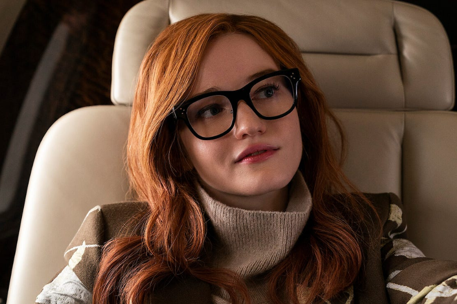 A woman with long red hair and large black-rimmed glasses gazes thoughtfully while seated in a cream-colored leather chair, wearing a taupe turtleneck sweater and a patterned coat. The soft lighting and luxurious surroundings suggest she is on a private jet.