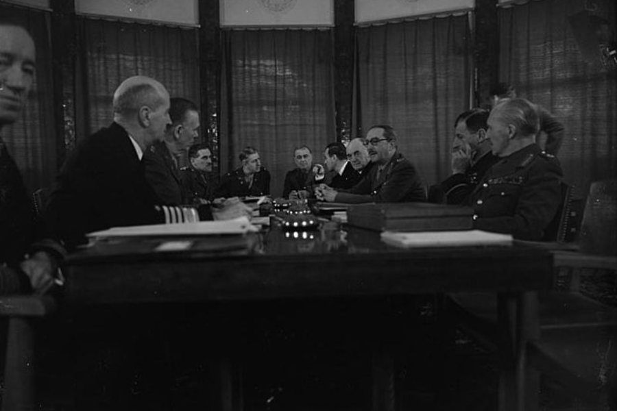 Black and white photo of a formal military meeting with high-ranking officers seated around a long table. The group includes naval and army personnel in uniform, engaged in discussion in a dimly lit room with curtained windows.