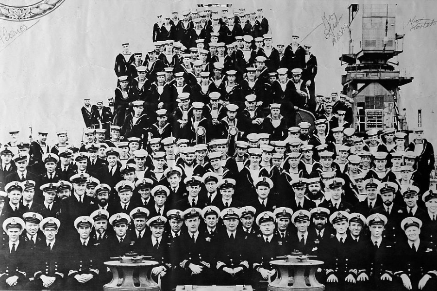 Black and white photo of a large group of naval personnel arranged in a pyramid formation aboard a ship. The crew members, all in uniform with caps, include officers and sailors, with some handwritten signatures visible on the photo.