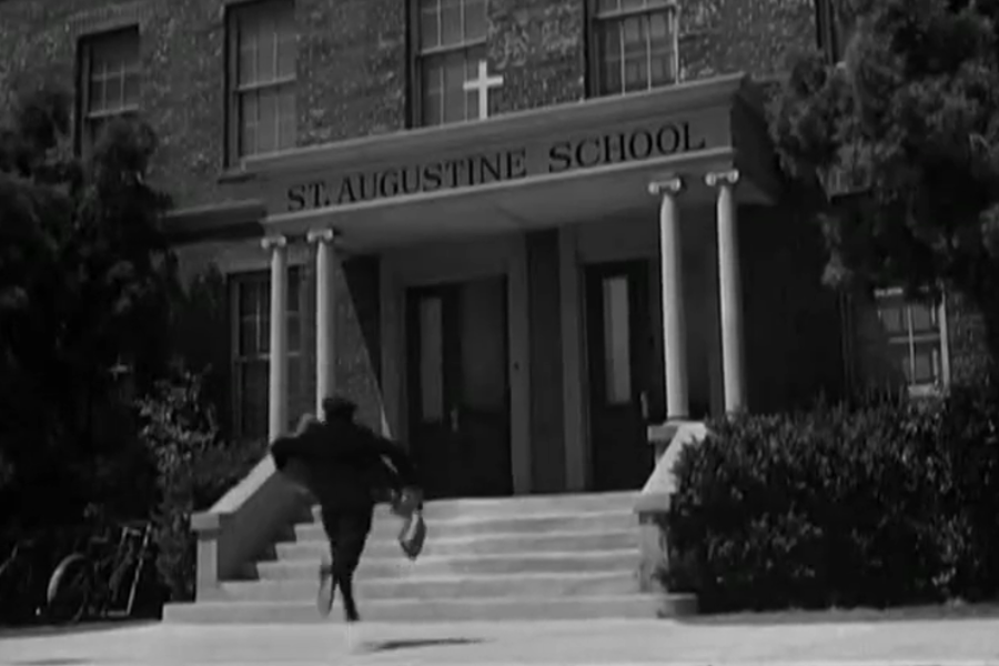 Black and white image of a person running up the steps toward the entrance of a building labeled “St. Augustine School.” The entrance features columns and a cross above the doorway, with trees and a bicycle partially visible on either side.