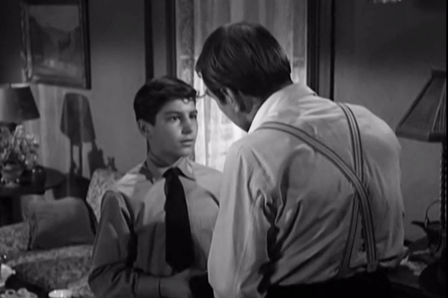 Black and white scene of a young boy in a dress shirt and tie standing in a modestly furnished living room, facing an older man in suspenders who appears to be speaking to him seriously. The room is softly lit with vintage décor and lace curtains in the background.