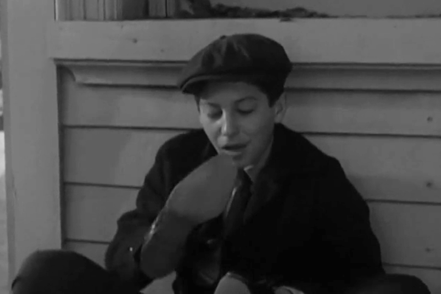 Black and white still of a young boy sitting on a porch or wooden stoop, dressed in a newsboy cap and dark coat. He’s looking down while adjusting or inspecting a shoe, with a calm, focused expression.