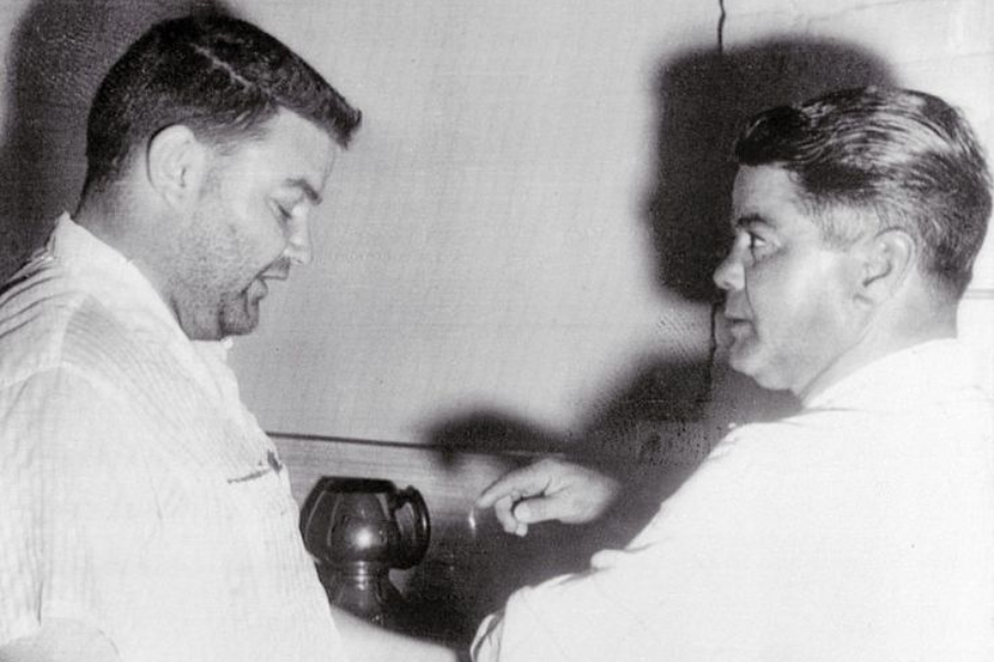 Black and white photo of two men in light-colored shirts, with one appearing to check or treat the other's arm in a casual setting. A pitcher or jug is seen in the background, and strong lighting casts distinct shadows on the wall.