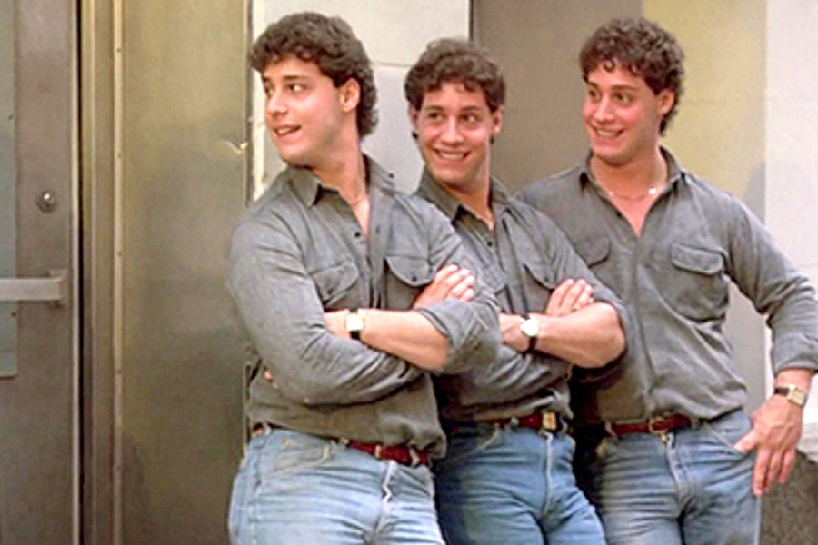 Three young men with identical features pose confidently outside a building, all wearing matching gray button-up shirts tucked into blue jeans with brown belts and watches. Their cheerful expressions and mirrored outfits highlight their striking resemblance as triplets.