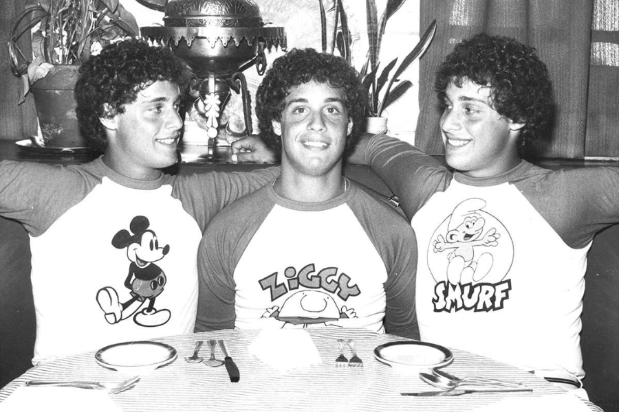 Black-and-white photo of three identical young men with curly hair sitting closely at a table, each wearing matching raglan shirts featuring cartoon characters: Mickey Mouse, Ziggy, and Smurf. They smile and lean into each other, exuding playful camaraderie.
