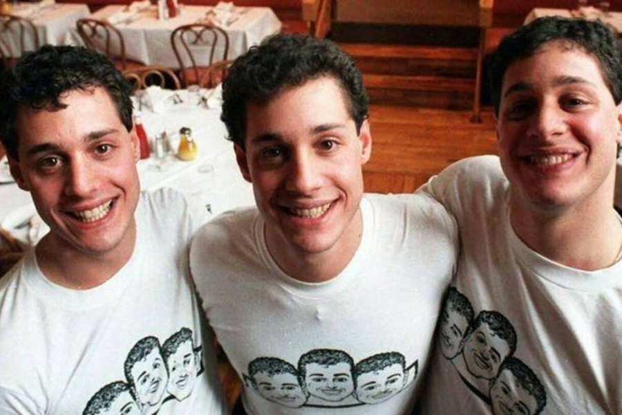 Three smiling young men with matching features stand arm-in-arm in a restaurant, all wearing identical white T-shirts printed with cartoon versions of their own faces. Their joyful expressions and coordinated outfits emphasize their identity as triplets.