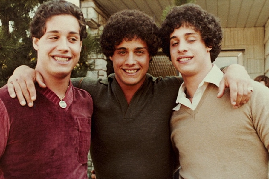 Three young men with identical features stand arm-in-arm outdoors, smiling and dressed in 1980s-style sweaters—one in burgundy, one in dark green, and one in tan. They appear close and happy, highlighting their bond as reunited triplets.