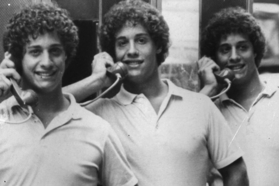 Black-and-white photo of three identical young men with curly hair smiling while each holds a phone receiver to their ear in a phone booth. All are dressed in light-colored polo shirts, emphasizing their shared features and playful energy.