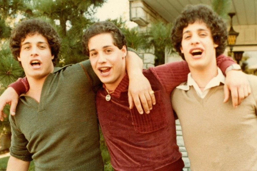Three young men with identical curly hair and facial features smile and laugh with their arms around each other outside, wearing 1980s-style sweaters. Their joyful expressions and close physical stance highlight their bond, suggesting they are triplets.