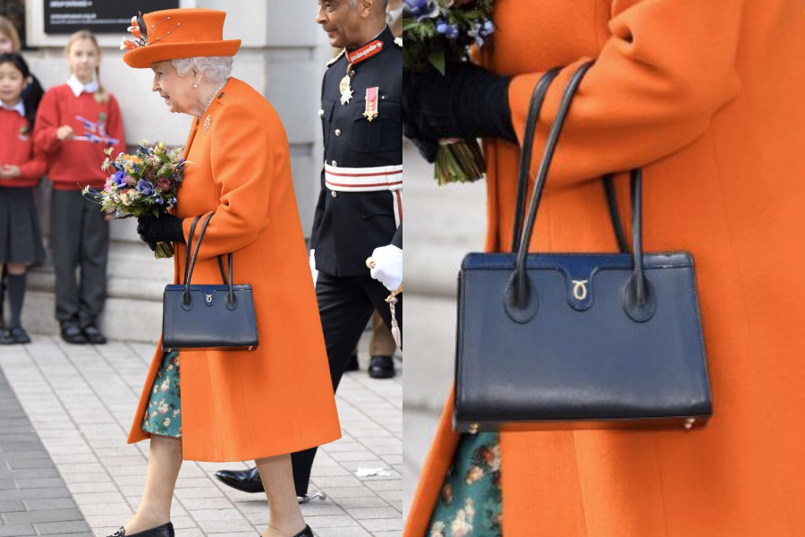 An elderly woman, dressed in a vibrant orange coat and matching hat adorned with a feather, carries a bouquet of flowers and a structured navy handbag. A close-up shows the handbag's smooth leather texture and gold hardware detail, with two top handles and a subtle emblem. She is interacting with school children and accompanied by a man in a formal military uniform.