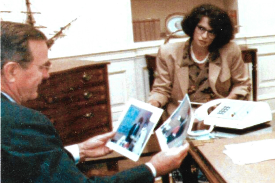 A man and woman sit across from each other in a formal office setting. The man, smiling, holds up two photographs, while the woman looks on seriously from behind a desk that holds a telephone, papers, and additional photos.