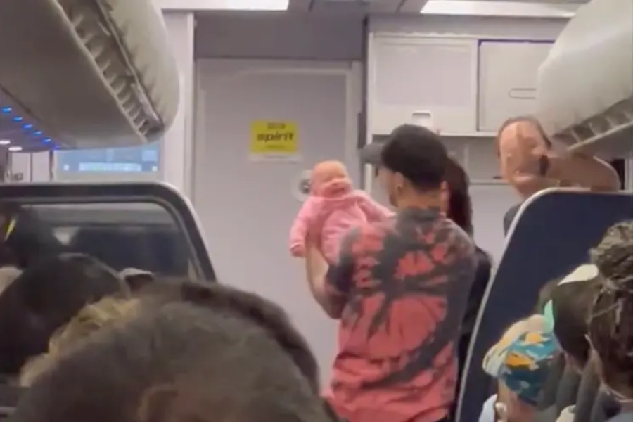 A man wearing a tie-dye shirt and cap holds up a smiling baby dressed in pink inside an airplane cabin. The Spirit Airlines logo is visible on the door in the background, with passengers seated nearby.