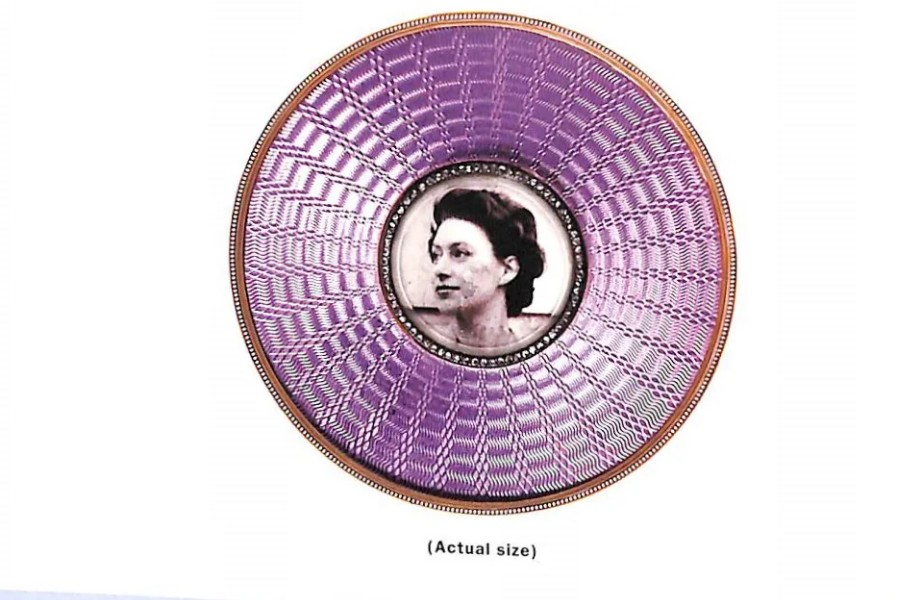 A small, circular portrait frame with an ornate guilloché enamel design in vivid purple radiating from the center, bordered with gold. At the center is a black-and-white photograph of a woman with dark hair styled in an elegant updo, encircled by a row of pearls or small crystals. The text beneath reads “(Actual size).”