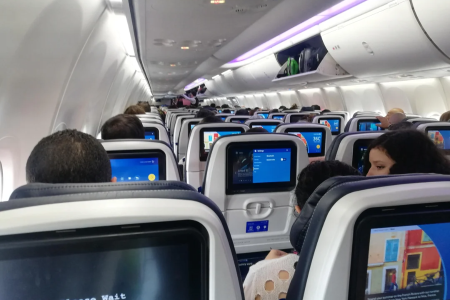 Interior of a modern airplane cabin with rows of passengers seated, each seat back equipped with a personal entertainment screen. Overhead bins are open with luggage inside, and soft purple lighting illuminates the cabin.
