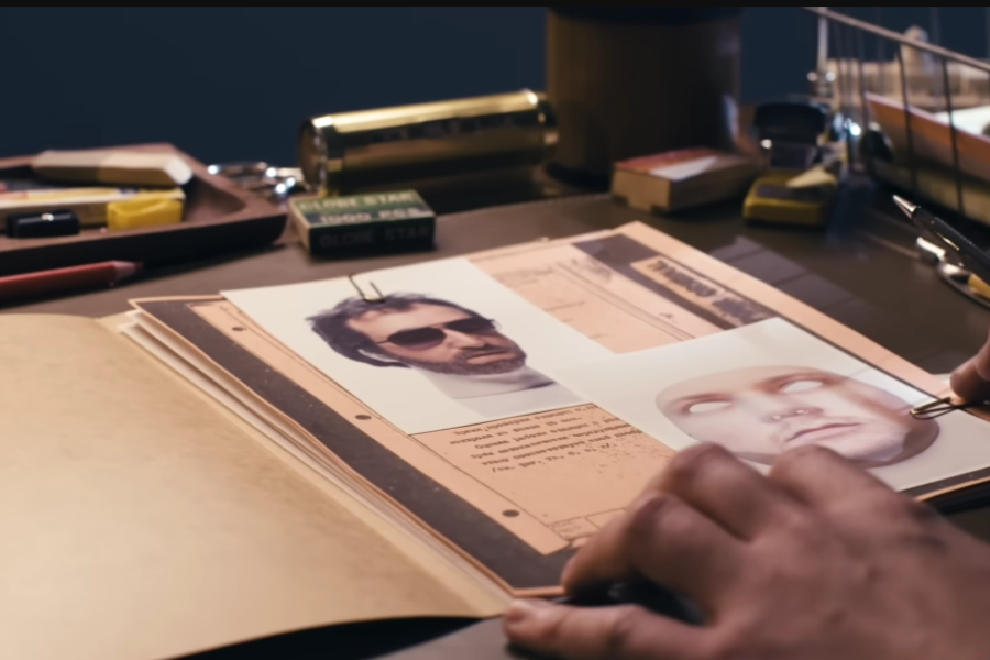 Close-up of a person's hands examining a dossier containing two photos—one of a man with sunglasses and another of a realistic face mask with blank white eyes. The desk is cluttered with office supplies, including a pencil, erasers, tape, and a label reading “GLOBE STAR.”