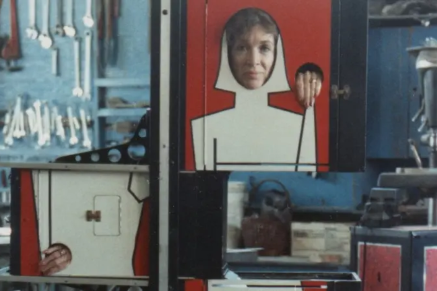 A woman poses inside a magician’s illusion box painted with a stylized female figure, with her face, hand, and foot emerging from separate compartments. The scene is set in a workshop with tools and machinery visible in the background.