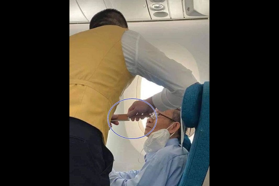 A flight attendant in a yellow vest helps an elderly passenger seated by the window drink from a cup using a straw. The passenger is wearing glasses and has a face mask pulled down below his chin.