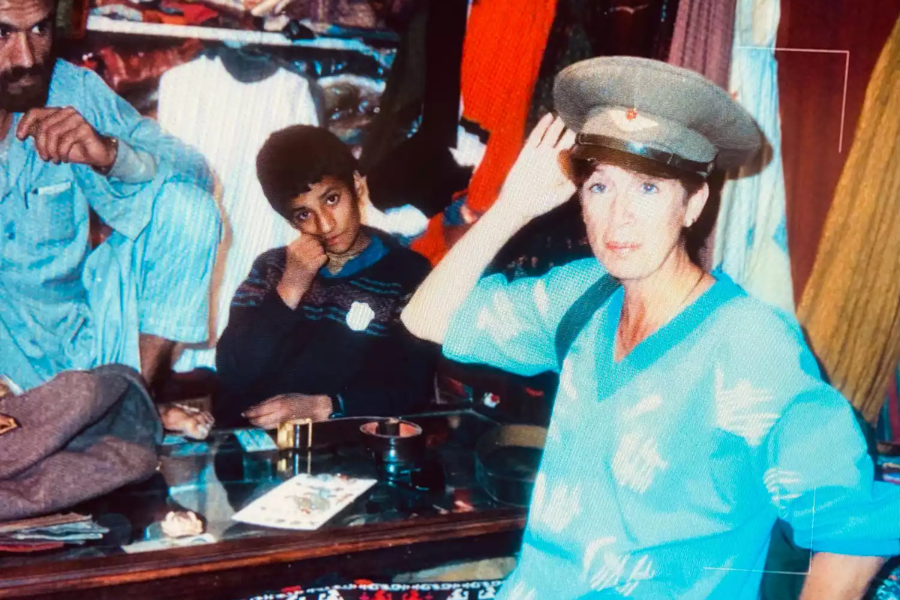 A woman in a blue sweater tries on a military-style hat inside a colorful shop, posing playfully while seated beside a young boy and a man dressed in traditional clothing. The shop is filled with textiles and various items displayed on a glass-topped counter.
