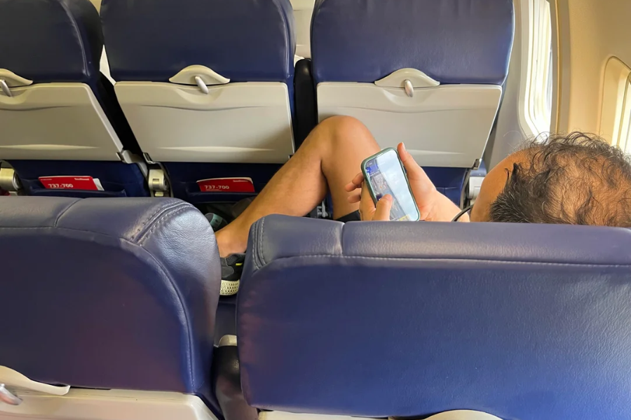 Passenger on an airplane is reclining across an empty row, with legs stretched out and watching something on a smartphone. The cabin seats are blue, and tray tables are folded up on the row in front.