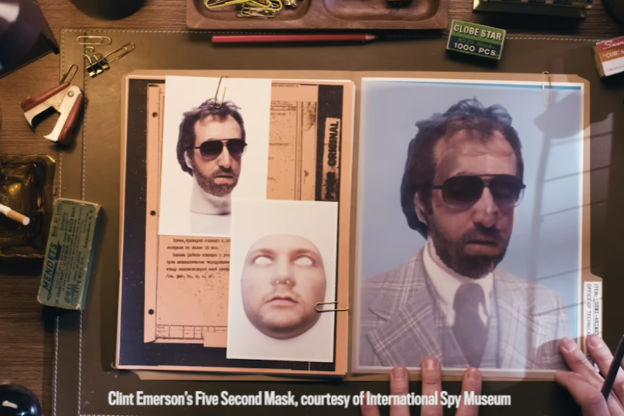 Overhead view of a desk displaying espionage materials, including a dossier with two printed disguise images—one of a man with sunglasses and another of a realistic face mask with blank eyes—next to a larger photo of a man in a plaid suit and sunglasses. The caption reads, “Clint Emerson’s Five Second Mask, courtesy of International Spy Museum.”