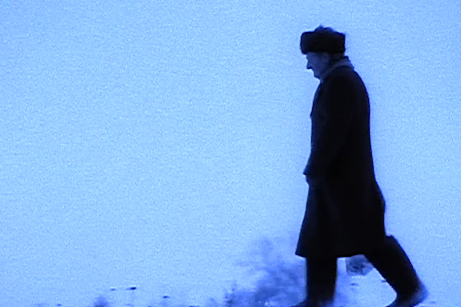 Silhouetted figure of a man in a long coat and fur hat walking alone through a snow-covered landscape under a cold blue hue. The scene evokes a somber, wintry atmosphere with minimal background detail.