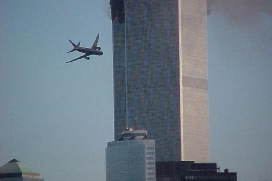 A commercial airplane is seen approaching one of the Twin Towers of the World Trade Center, with dark smoke and fire visible from the upper floors of the building. The scene captures a moment during the tragic events of September 11, 2001.