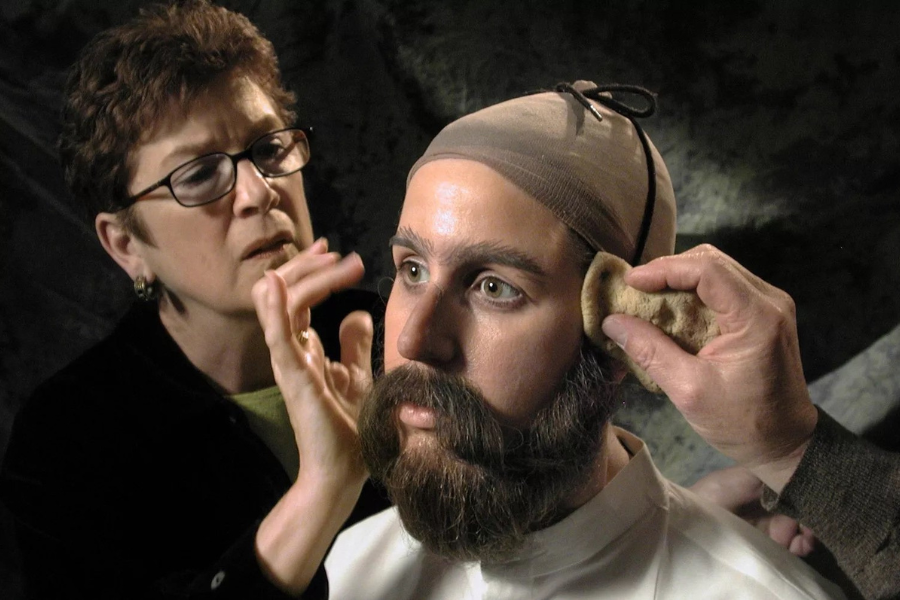 Two people work on applying a hyper-realistic disguise to a mannequin or person, attaching a fake beard and makeup while adjusting facial details. The individual being transformed wears a beige wig cap and has strikingly lifelike features, showcasing special effects or spy disguise craftsmanship.