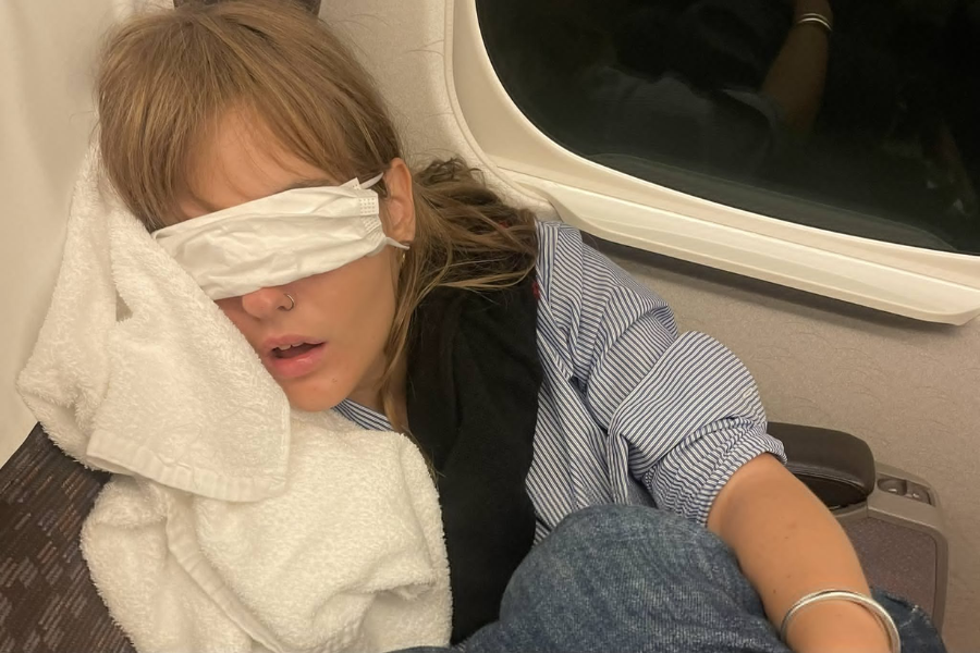 A passenger is asleep on an airplane seat, resting their head on a towel and using a face mask as an improvised sleep mask. The person appears relaxed, with a window visible in the background.