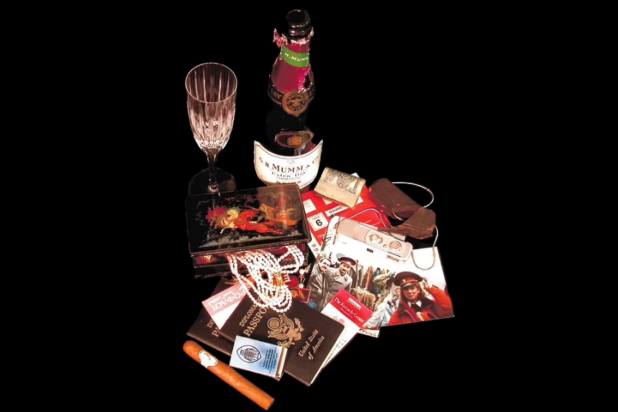 Collection of espionage-themed items arranged against a black background, including a champagne bottle and glass, fake passports, pearl necklaces, cash, a cigar, a cassette tape recorder, tourist brochures, and sunglasses. The display suggests a spy’s cover identity or travel kit.