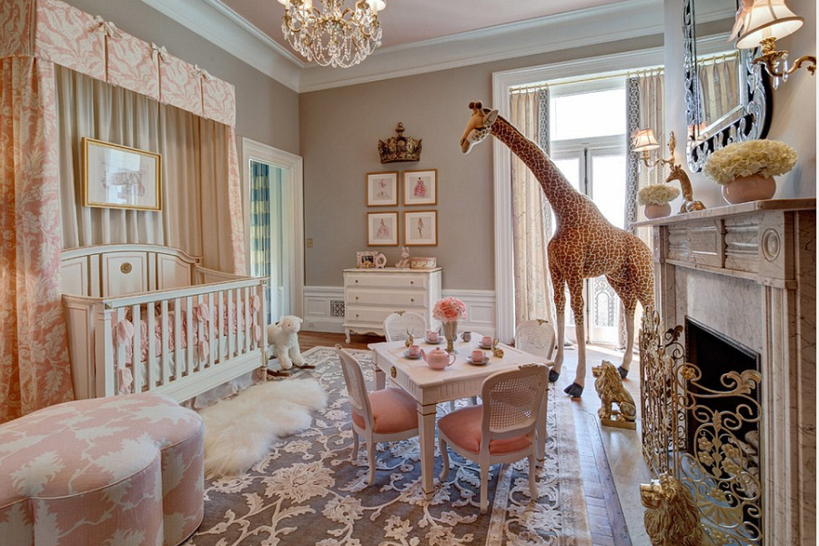 A lavishly decorated nursery featuring a soft pink and ivory color scheme, with a white crib draped in floral curtains, a tea table set for four with pink chairs, and a plush area rug. A life-size stuffed giraffe stands near the window, and the room is adorned with elegant touches including a crystal chandelier, floral arrangements, and a white marble fireplace with gold lion accents.