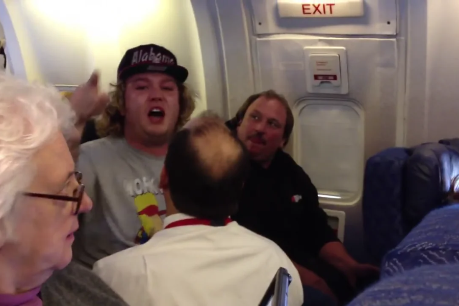 A man wearing an "Alabama" cap appears to be shouting or arguing near the exit door of an airplane, as other passengers and a crew member look on. The scene looks tense, with people seated nearby observing the situation.