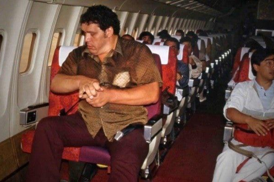 A large man sits on an airplane, occupying part of two seats, as he adjusts his seatbelt. The cabin has red patterned seats, and other passengers are visible seated in the background.