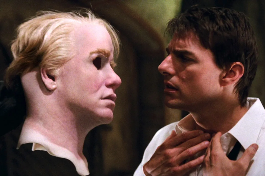 A tense scene featuring a man pulling at his collar while staring at a hyper-realistic silicone mask of a woman’s face, which is hollow at the eyes and head. The dramatic lighting and expressions suggest a moment of revelation or disguise in a spy or action film.